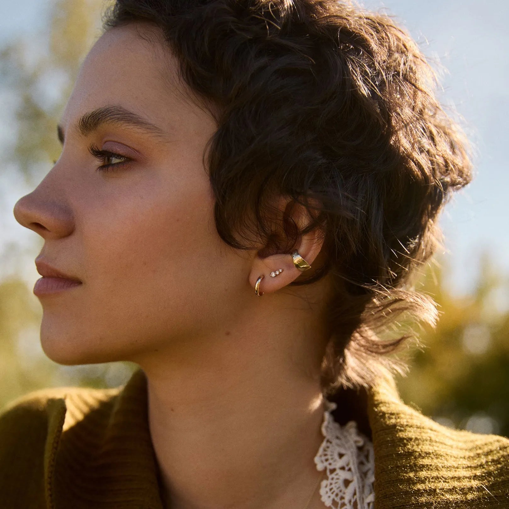 Woman wearing a brown sweater with lace trim, standing outdoors with trees in the background.