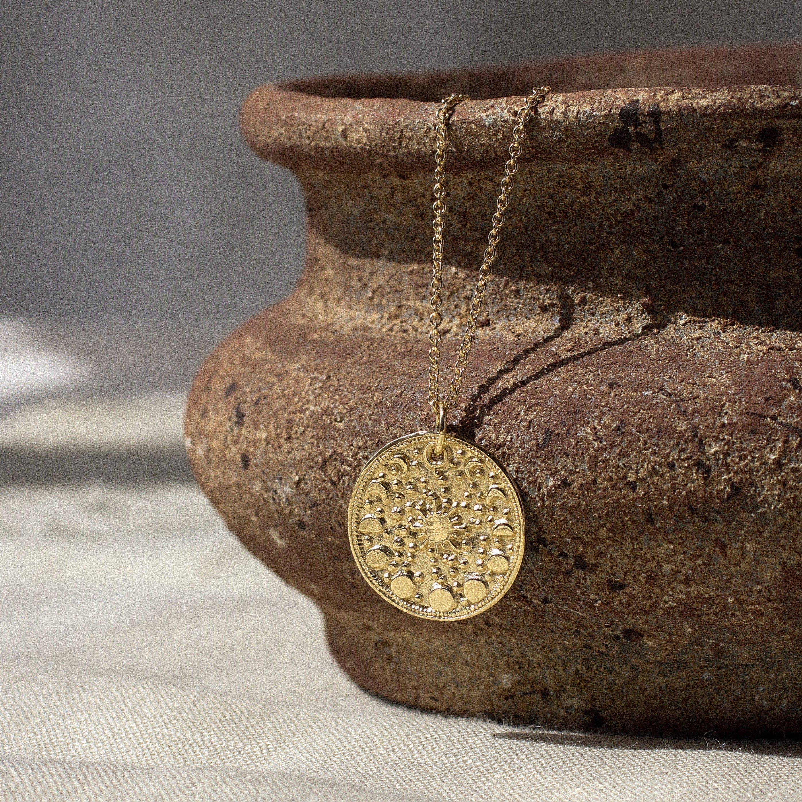 gold moonphase round pendant necklace hanging on a ceramic dish with shadows on a fabric surface