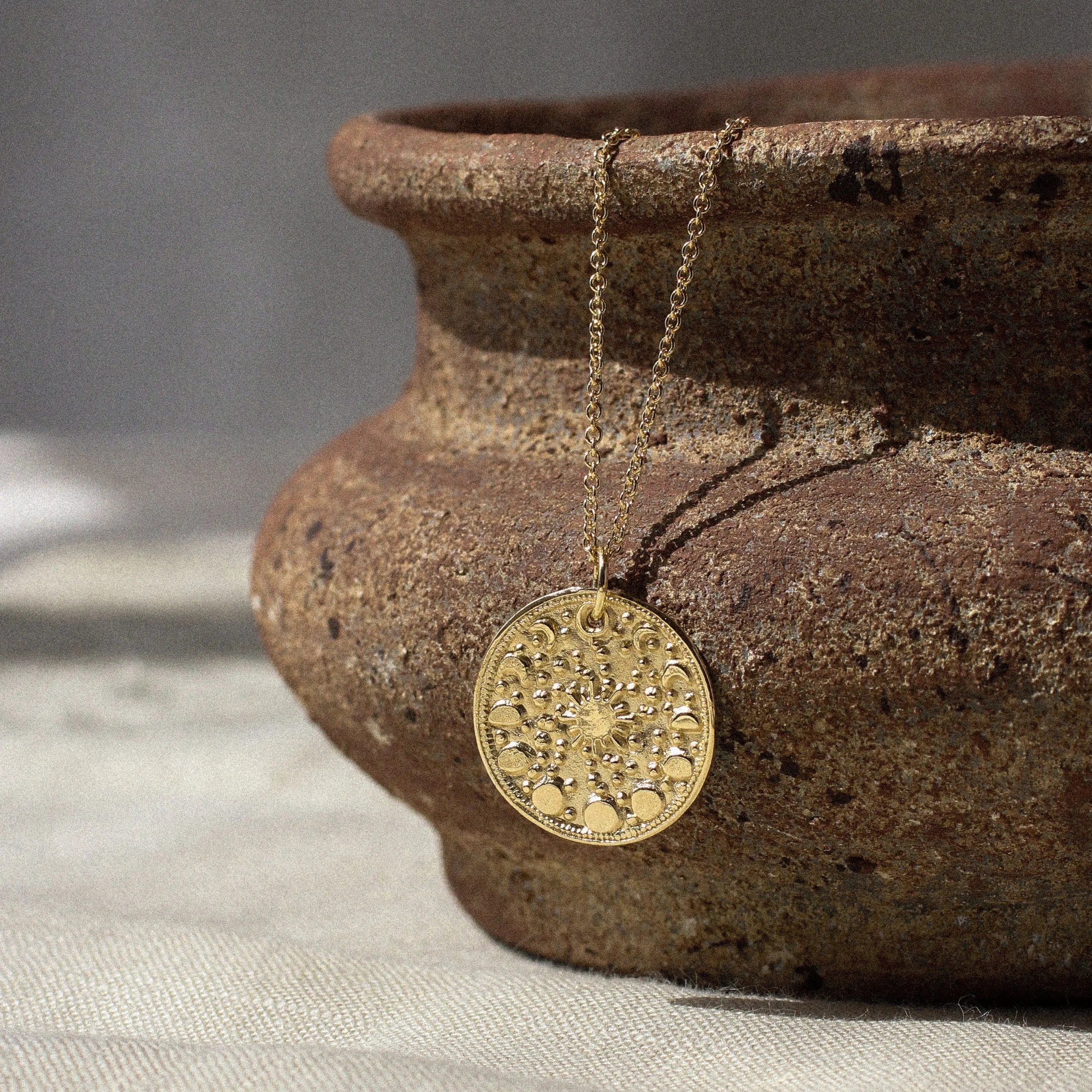 gold moonphase round pendant necklace hanging on a ceramic dish with shadows on a fabric surface