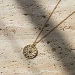 A gold-toned necklace with a textured circular pendant lies on a light beige stone surface under dappled sunlight and shadows.