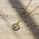A gold-toned necklace with a textured circular pendant lies on a light beige stone surface under dappled sunlight and shadows.