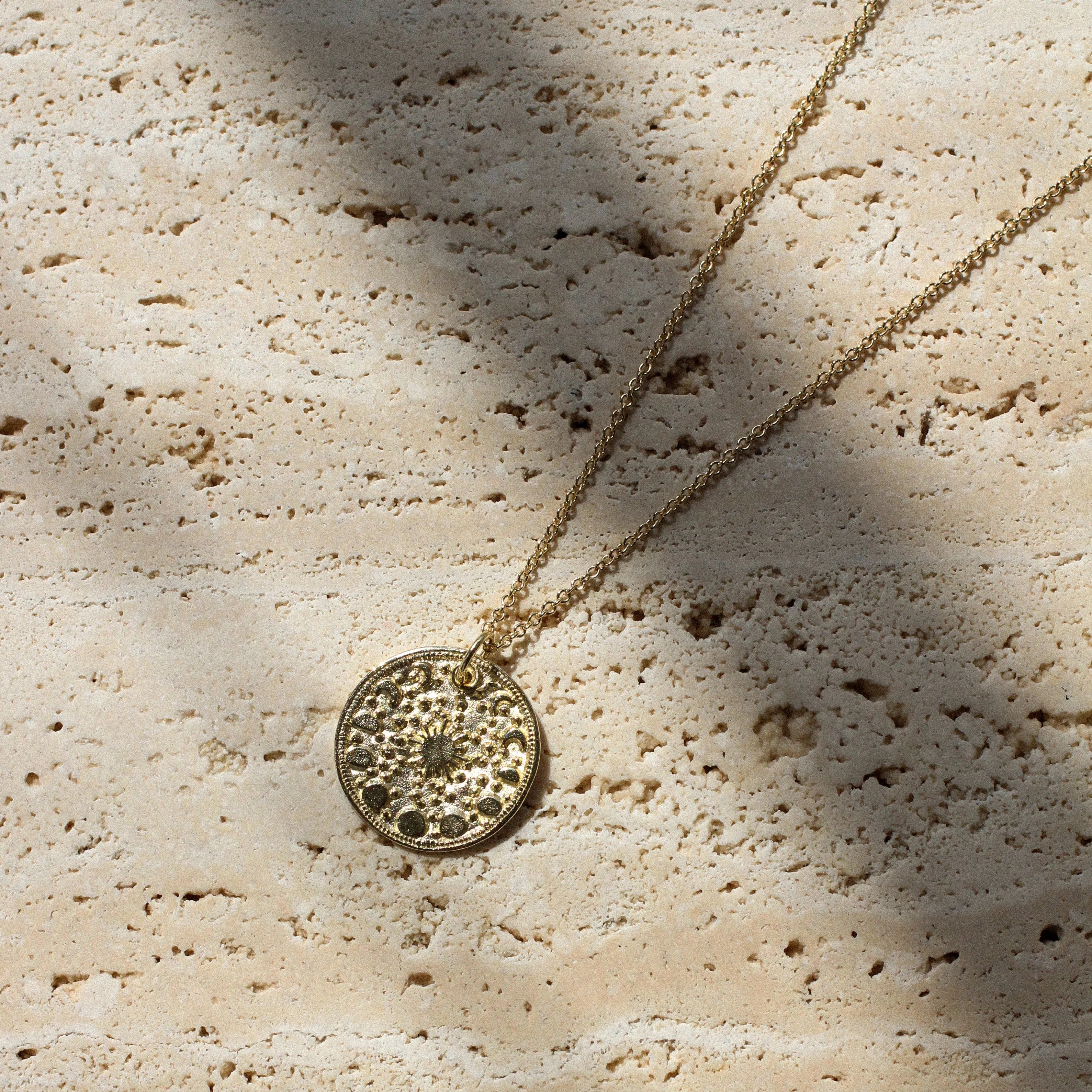 A gold-toned necklace with a textured circular pendant lies on a light beige stone surface under dappled sunlight and shadows.