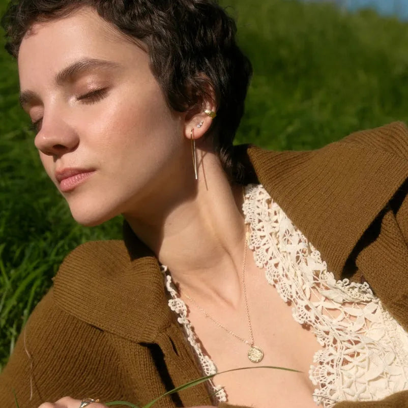 A woman with short curly dark hair lies in a grassy field under a blue sky, wearing a brown cardigan over a cream lace-trim top.
