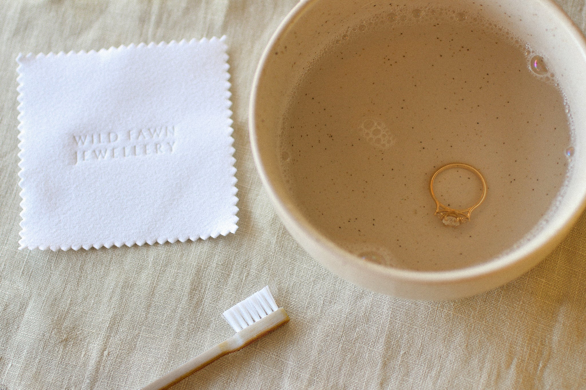 Gold ring in a bowl of soapy water with a cloth and toothbrush on a wooden surface