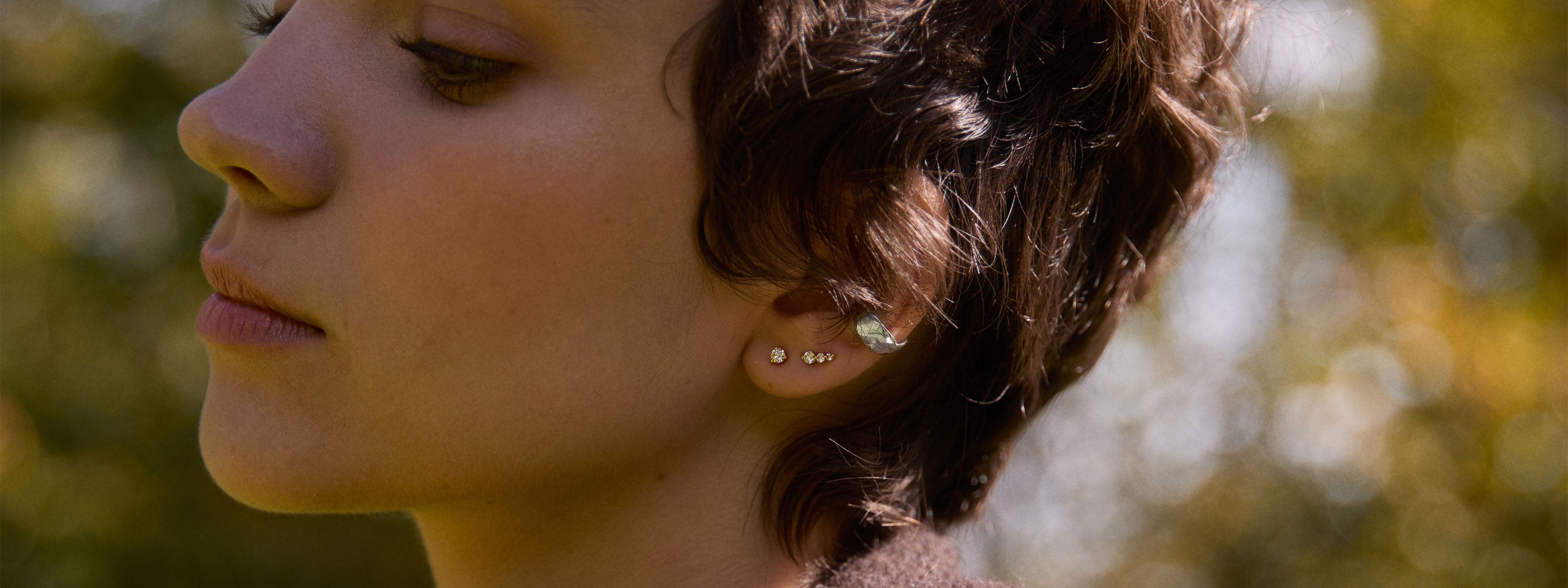 Close-up of a woman's face wearing diamond stud earrings and silver ear cuff with a blurred natural background