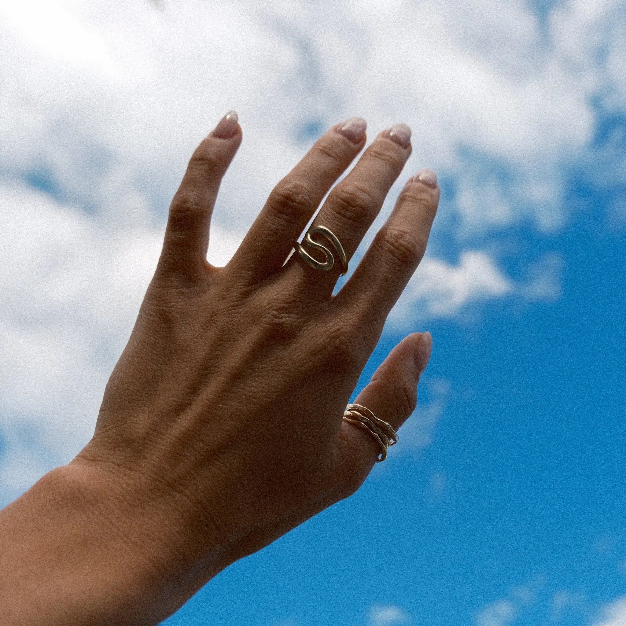 A hand with manicured nails wearing two gold rings, one of which is a twisted s-shaped band, set against a bright blue sky with white clouds.