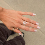 A close-up of a person's hand wearing a silver s-shaped ring on the ring finger, with a gold ring on the thumb, set against a neutral concrete background.
