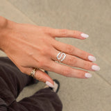 A close-up of a person's hand wearing a silver s-shaped ring on the ring finger, with a gold ring on the thumb, set against a neutral concrete background.