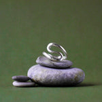 A silver ring with a sculptural, flowing S-shaped curve, displayed on a stack of smooth gray zen stones against a solid olive green background.