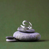 A silver ring with a sculptural, flowing S-shaped curve, displayed on a stack of smooth gray zen stones against a solid olive green background.