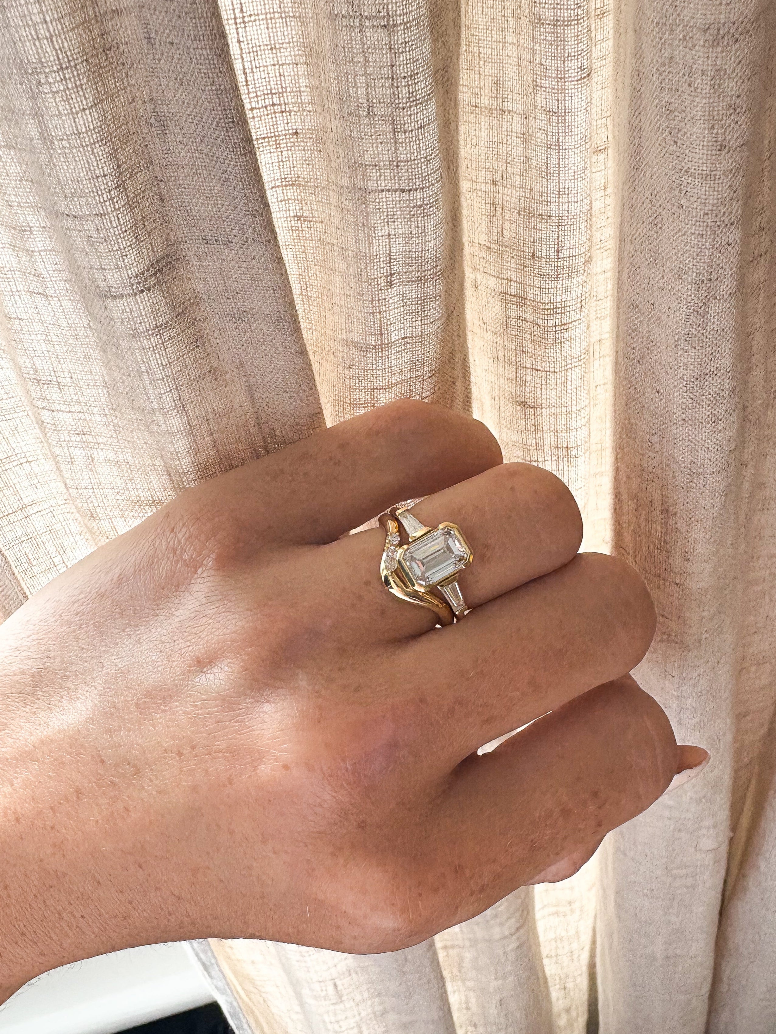 Hand wearing a gold ring with a diamond, set against a beige curtain background