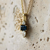A gold pendant necklace featuring a rectangular dark blue sapphire center stone, a small clear round diamond above it, and a pear-cut clear diamond below, displayed against a textured beige stone background.