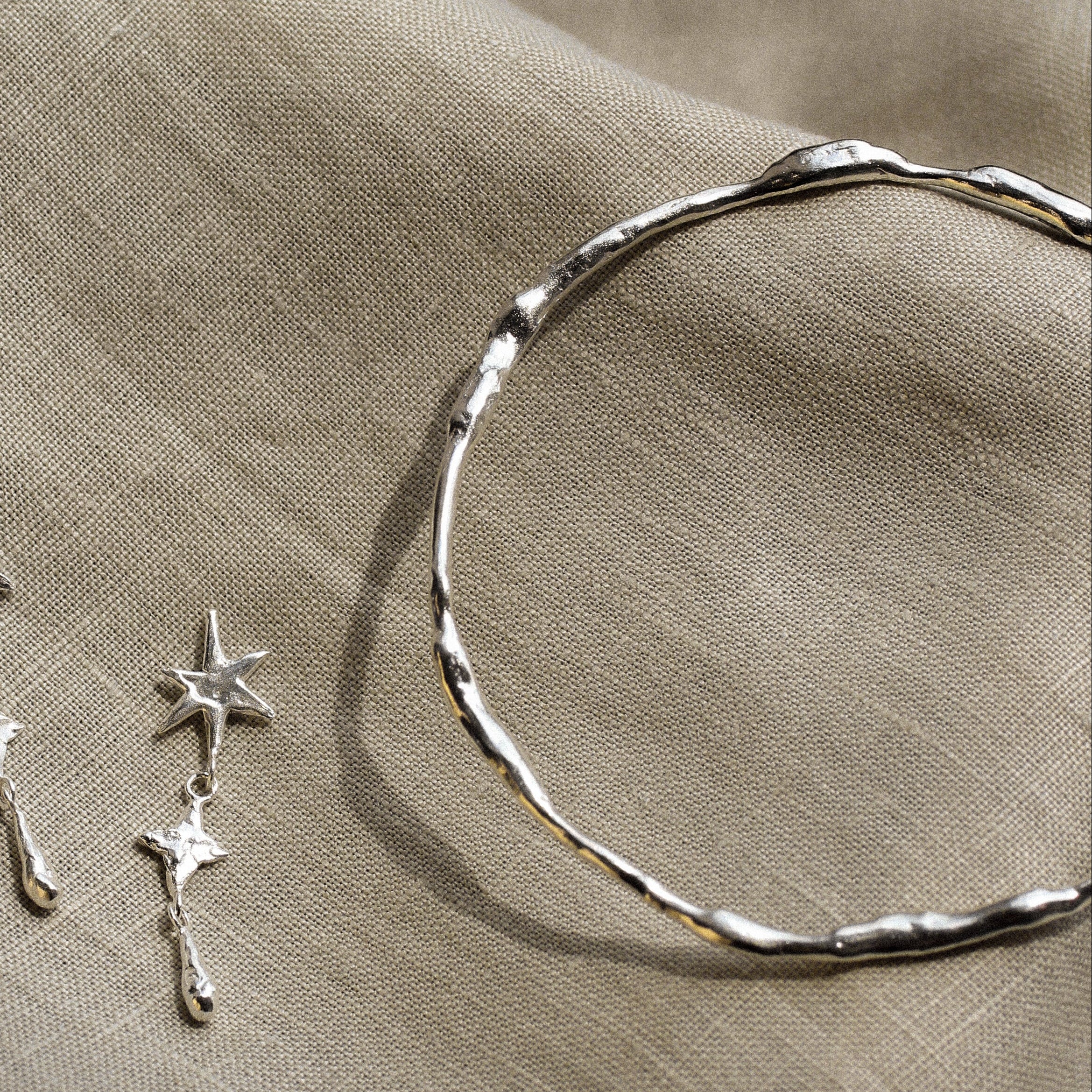 close up of silver organic bangle and a silver star dangle earring on a beige fabric
