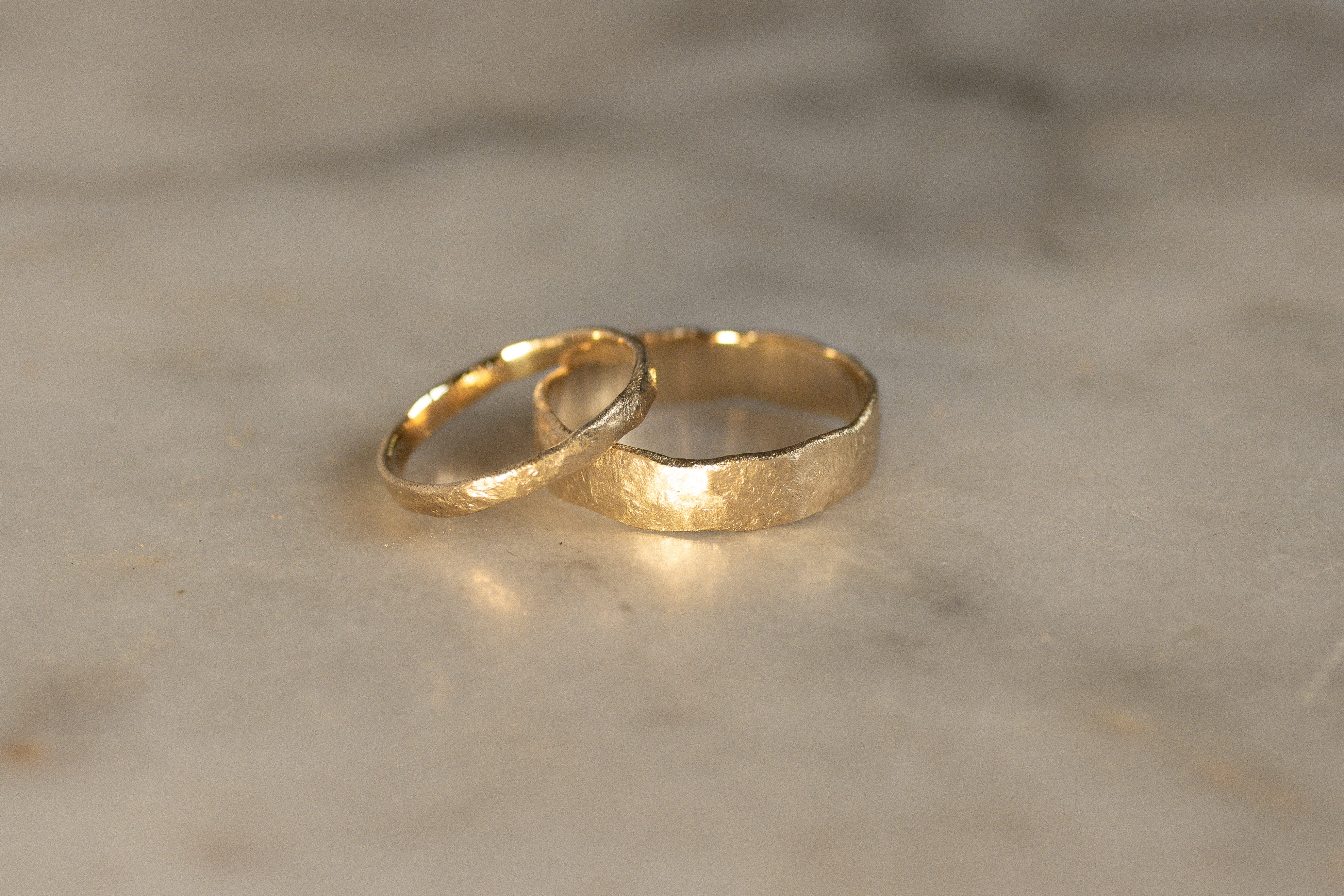 a 2mm wide and 5mm wide wedding ring in gold with uneven textures on a marble surface