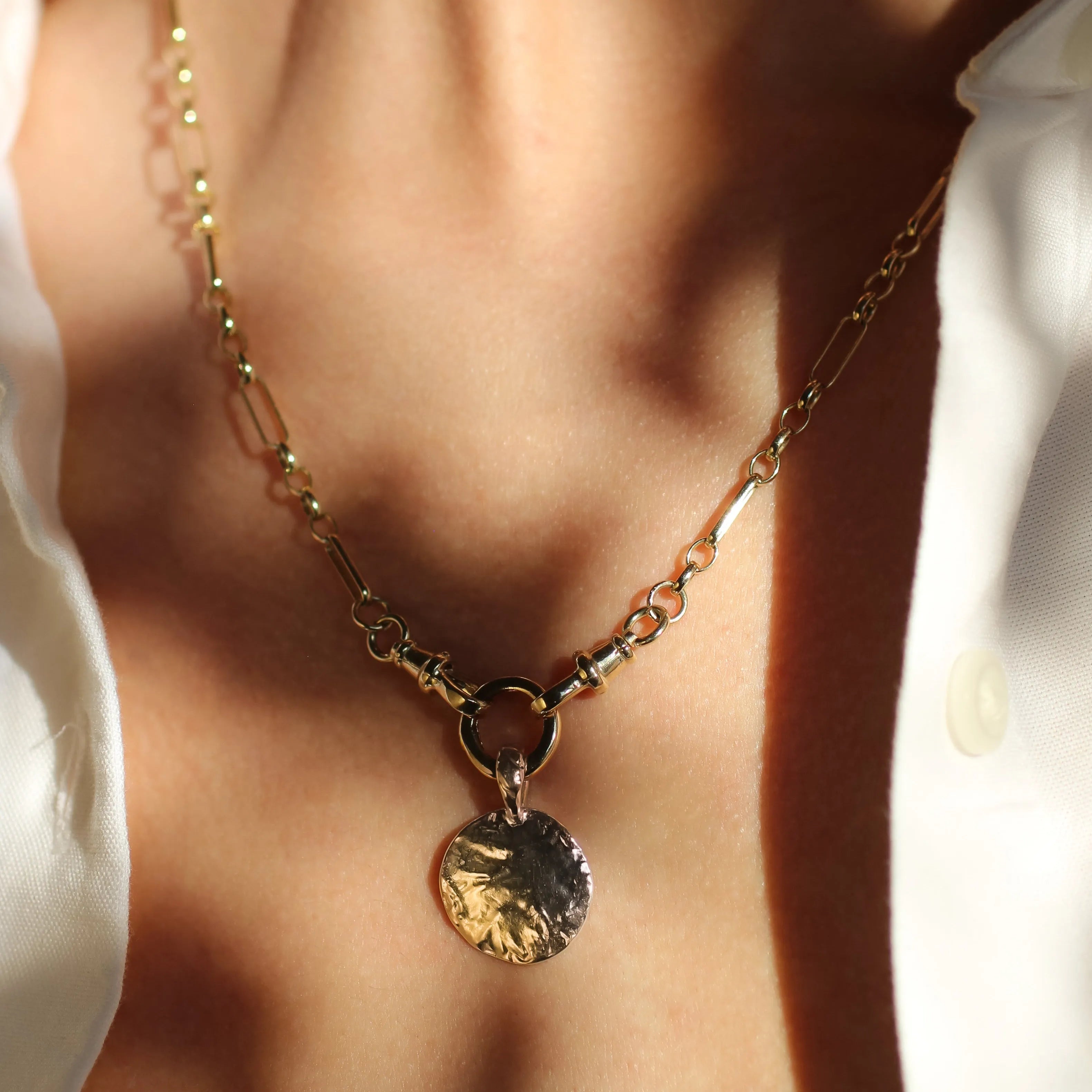 chunky gold necklace with a textured round pendant worn by a person in a white shirt, with shadows