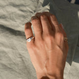 A close-up of a person's hand resting on a textured gray fabric surface, wearing a silver ring with a heart-shaped face on their little finger and a thin gold band on their thumb