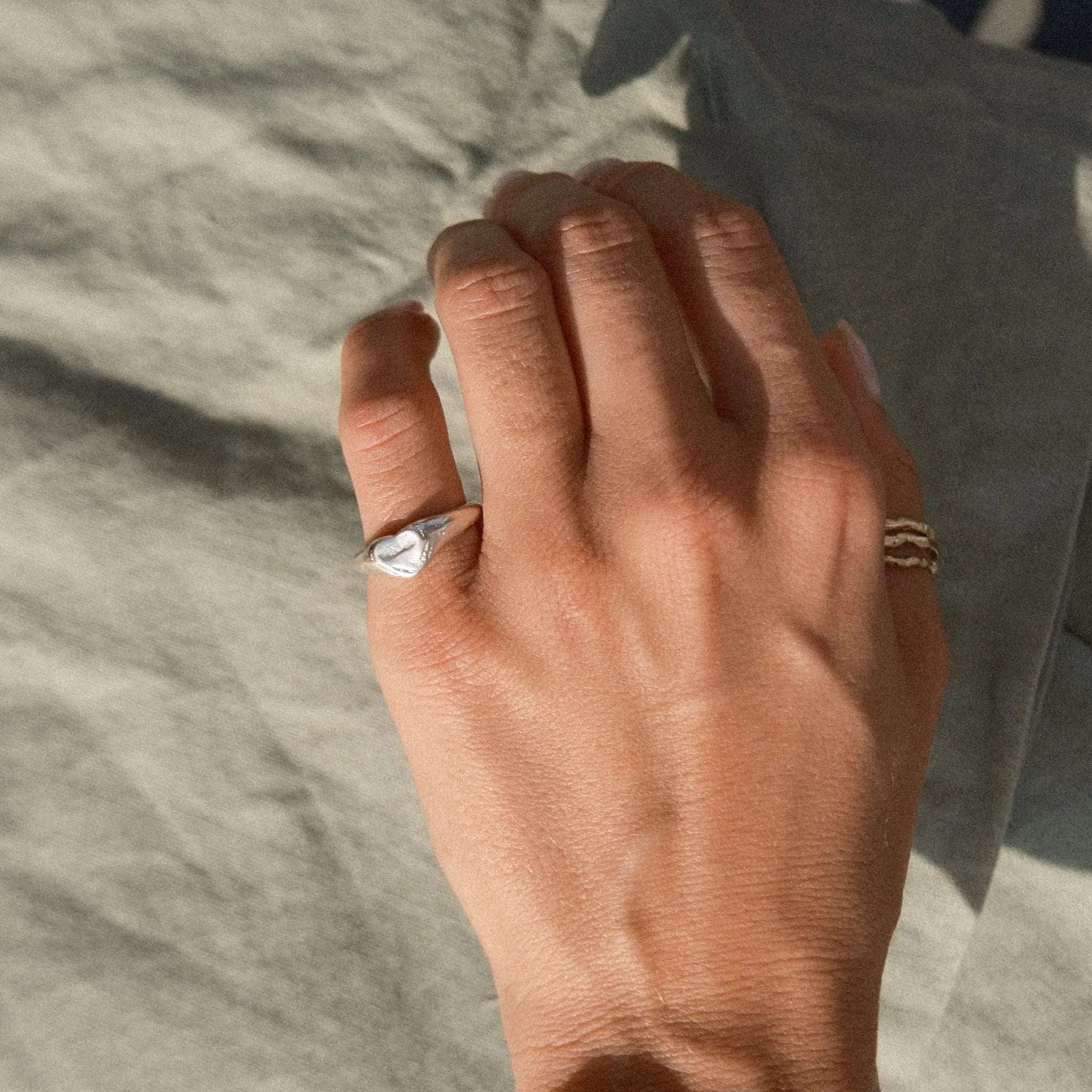 A close-up of a person's hand resting on a textured gray fabric surface, wearing a silver ring with a heart-shaped face on their little finger and a thin gold band on their thumb
