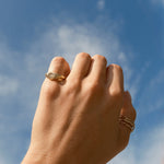 A close-up of a person's hand wearing a gold pinky ring with a textured, heart-shaped face, set against a bright blue sky with light clouds.