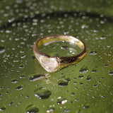 A gold ring with a heart-shaped clear stone, resting on a wet green surface covered in water droplets.