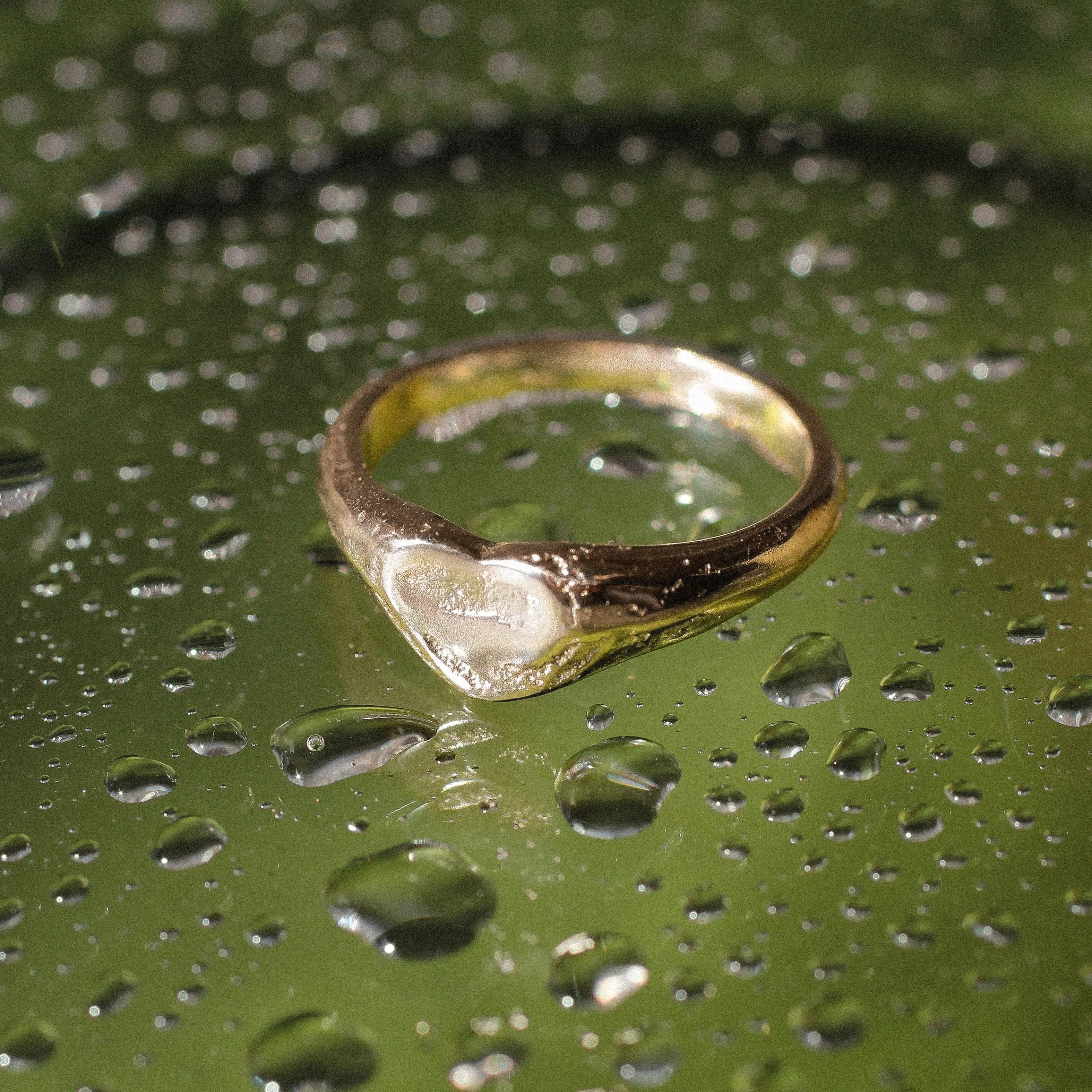 A gold ring with a heart-shaped clear stone, resting on a wet green surface covered in water droplets.