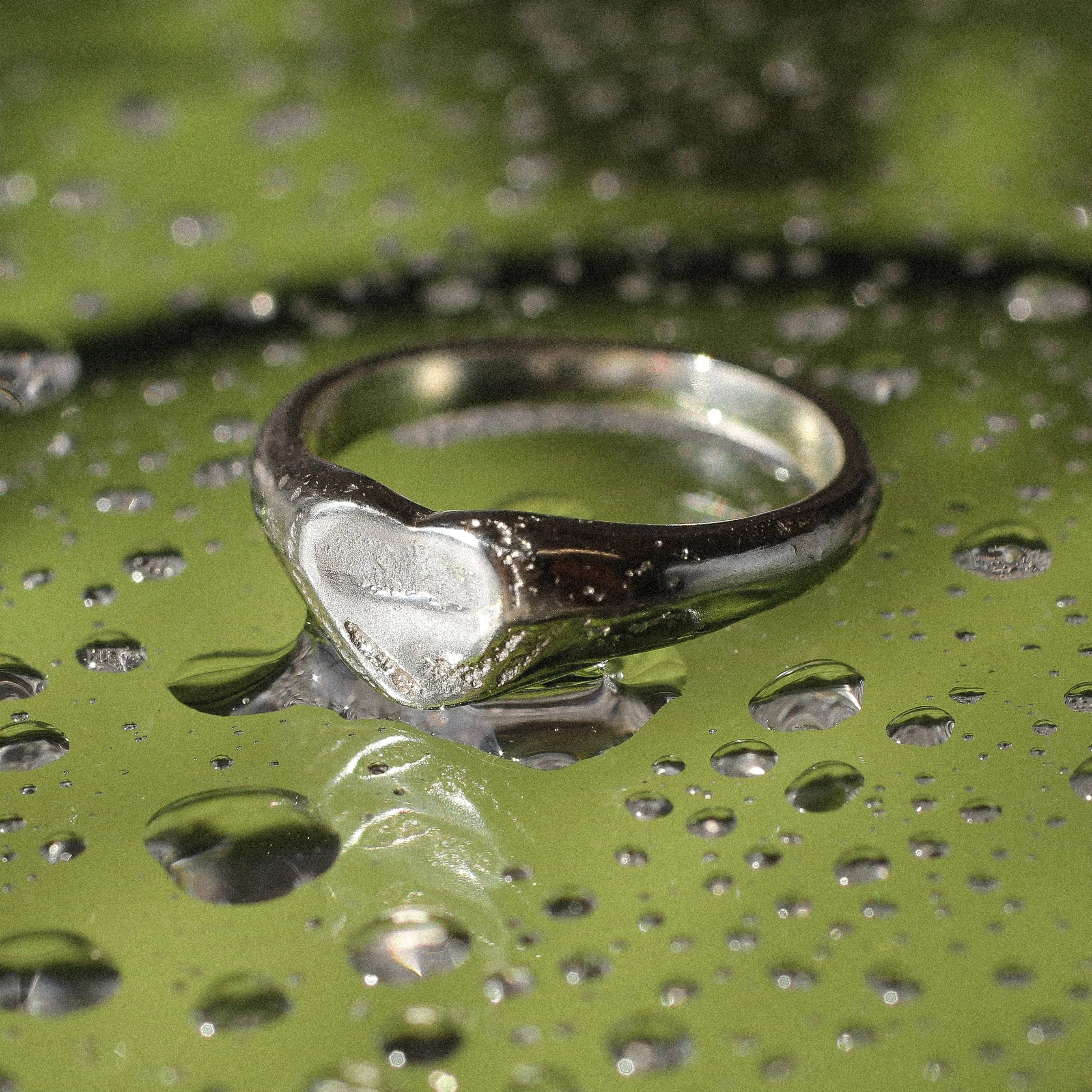 silver pinkie signet ring with heart-shaped design on a green surface with water droplets on it