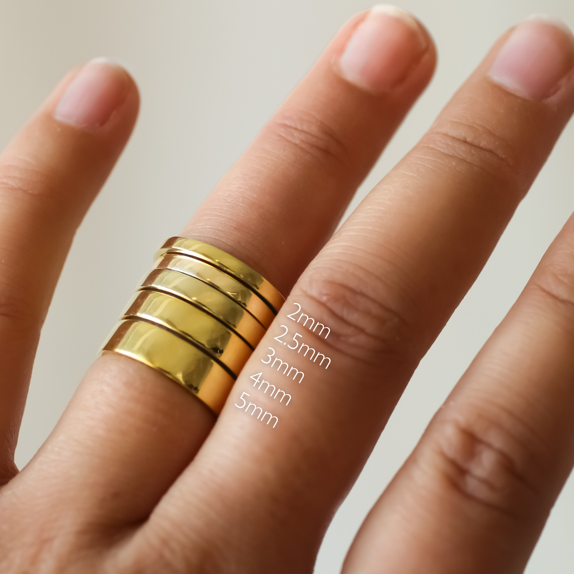 flat gold wedding rings on a finger with size markings 2mm 2.5mm 3mm 4mm and 5mm against a neutral background