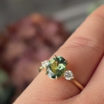 video of a greeny blue sapphire engagement ring on someones hand sparkling in the light, with some blurred pink flowers in the background