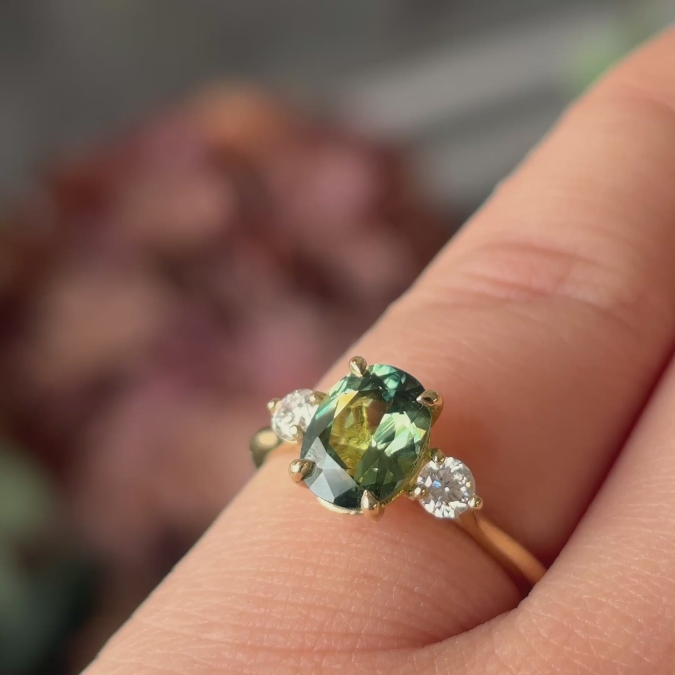video of a greeny blue sapphire engagement ring on someones hand sparkling in the light, with some blurred pink flowers in the background