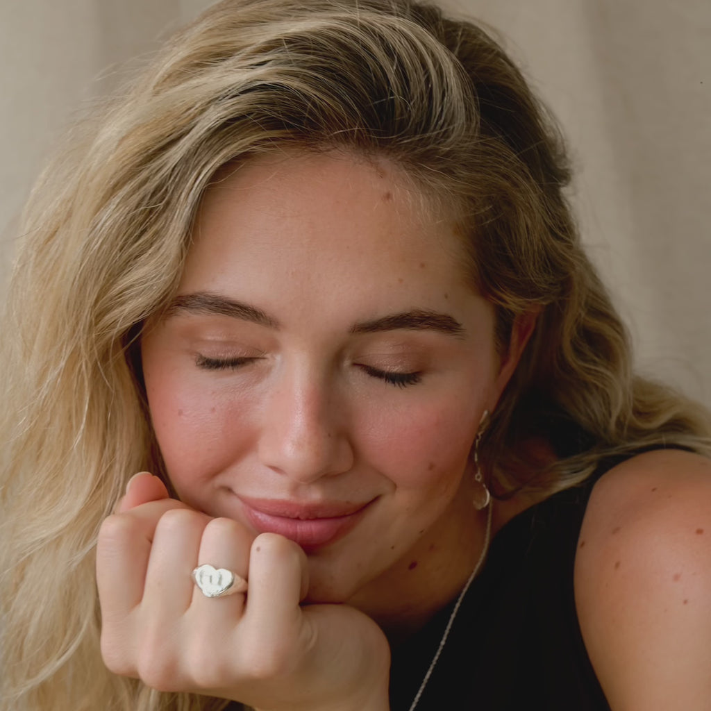 blonde model with long wavy hair resting her chin on her hand wearing a heart-shaped signet ring