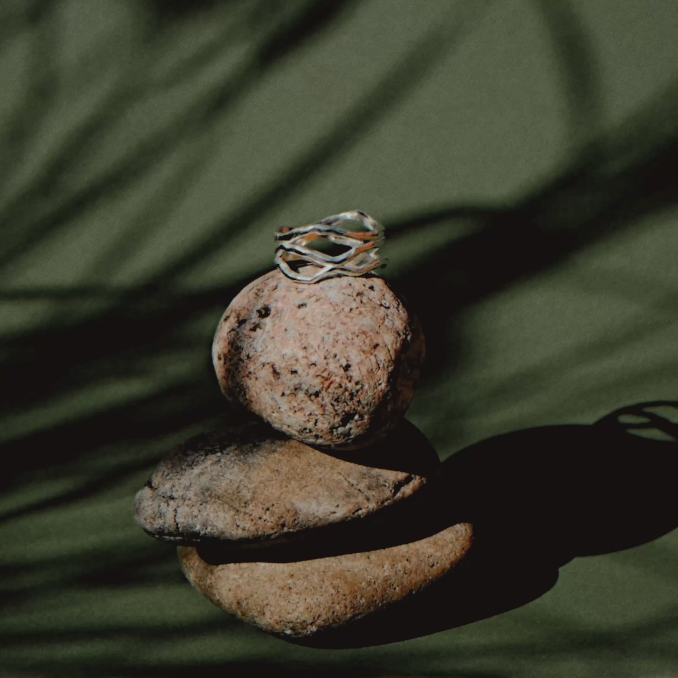 silver entwined ring on a rock stack against a green background with shadows of moving plants