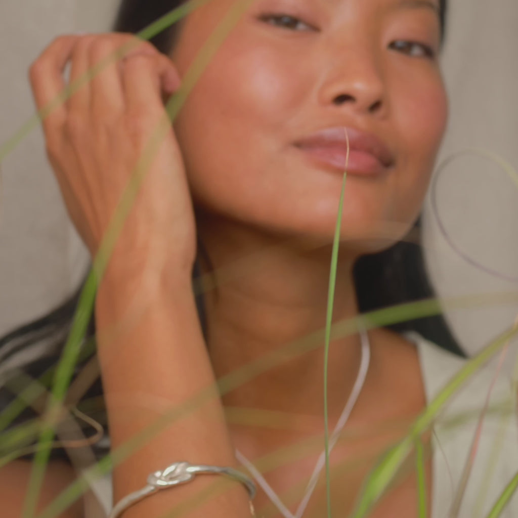 model tucking her hair behind her ear and leaning on her hand, wearing silver jewellery including knotted bangle and round studs