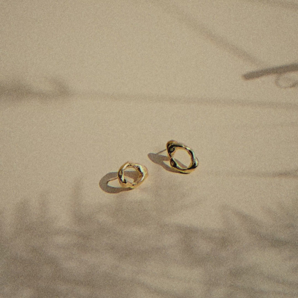 gold oval studs with molten texture on a beige surface with moving plant shadows