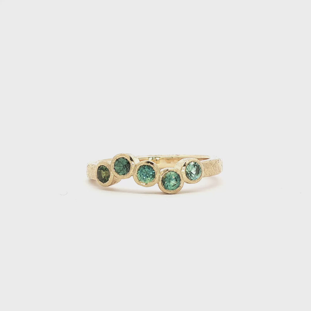 gold engagement ring with five round blue and green sapphires and textured band spinning on a turntable