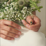 hands putting on a green sapphire engagement ring with tapered diamonds and flowers in the background