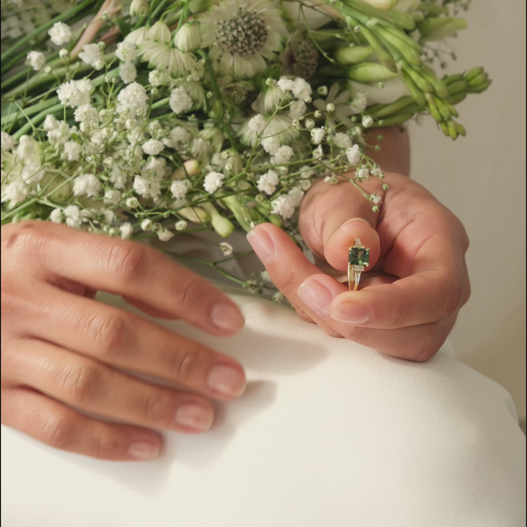 hands putting on a green sapphire engagement ring with tapered diamonds and flowers in the background
