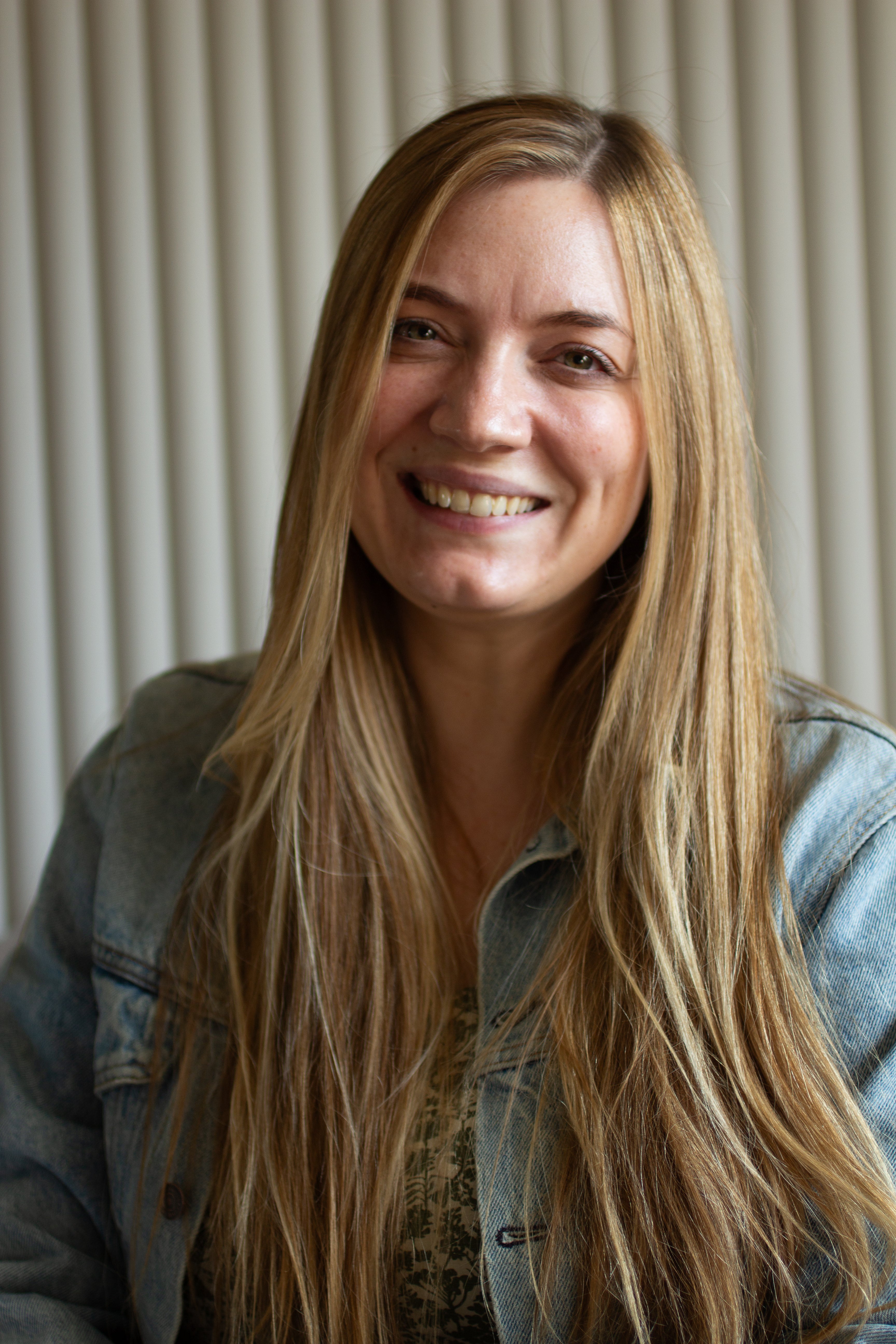 Woman with long blonde hair wearing a denim jacket against a neutral background