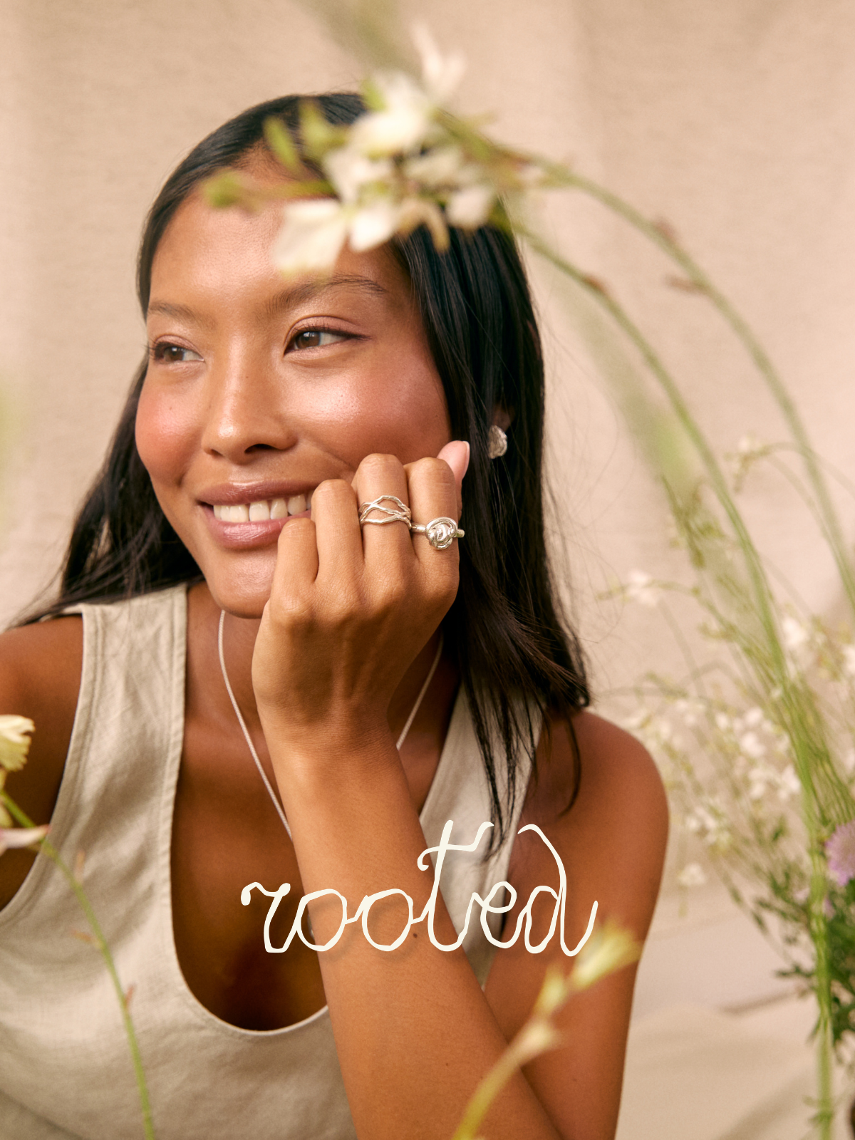 A woman with long dark hair smiles while wearing multiple silver rings and a delicate silver necklace. She is wearing a light beige tank top and posing with soft white flowers against a neutral beige background.