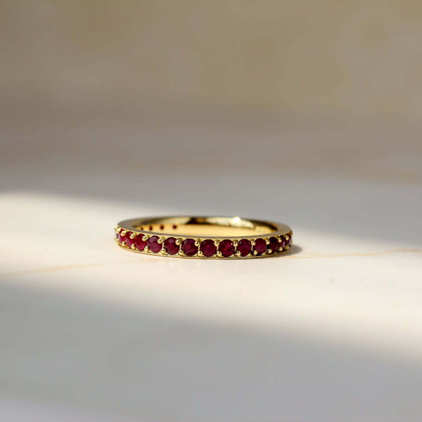A gold eternity ring set with small round ruby gemstones, resting on a light beige marble surface with soft natural shadows.