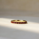 A gold eternity ring set with small round ruby gemstones, resting on a light beige marble surface with soft natural shadows.