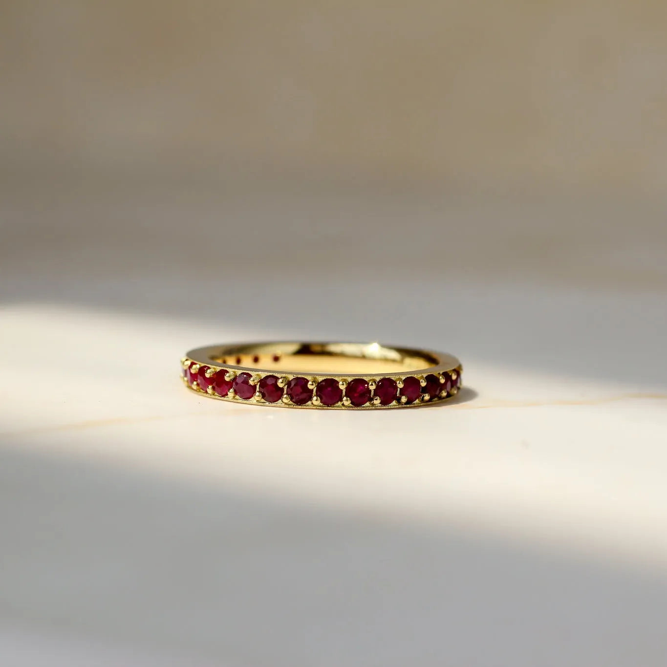 A gold eternity ring set with small round ruby gemstones, resting on a light beige marble surface with soft natural shadows.
