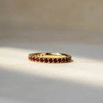 A gold eternity ring set with small round ruby gemstones, resting on a light beige marble surface with soft natural shadows.