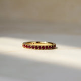 A gold eternity ring set with small round ruby gemstones, resting on a light beige marble surface with soft natural shadows.