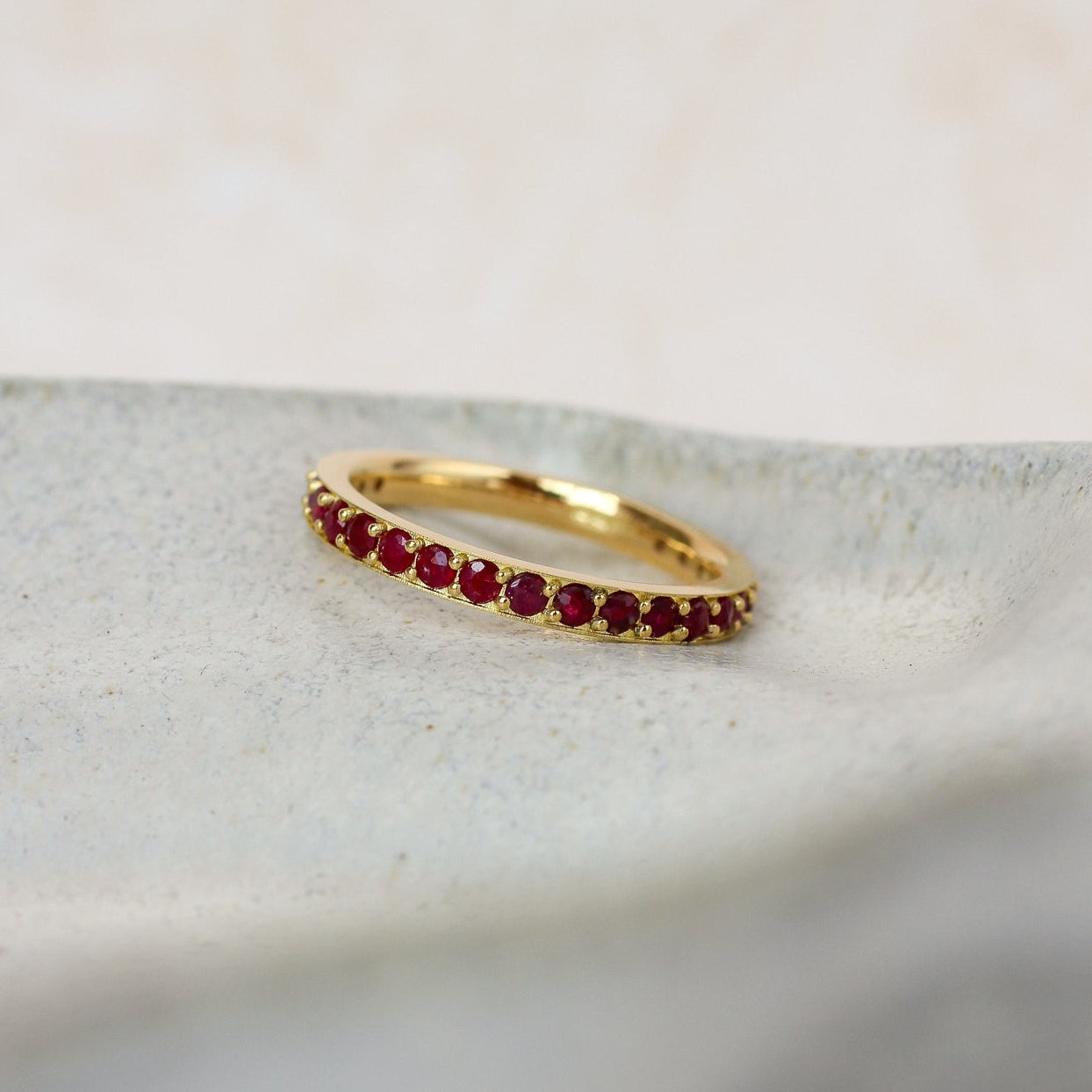 A delicate yellow gold ruby eternity ring featuring a row of small, round-cut deep red gemstones, resting on a textured light gray stone slab.