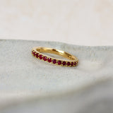 A delicate yellow gold ruby eternity ring featuring a row of small, round-cut deep red gemstones, resting on a textured light gray stone slab.
