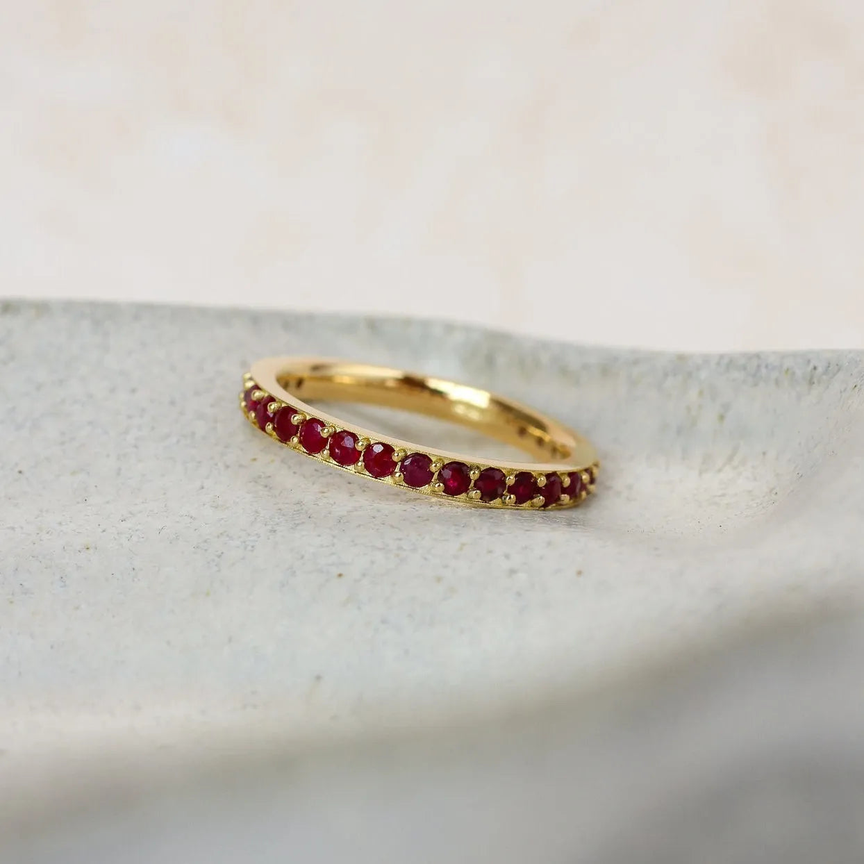 A delicate yellow gold ruby eternity ring featuring a row of small, round-cut deep red gemstones, resting on a textured light gray stone slab.