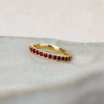 A delicate yellow gold ruby eternity ring featuring a row of small, round-cut deep red gemstones, resting on a textured light gray stone slab.