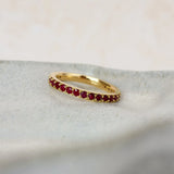 A delicate yellow gold ruby eternity ring featuring a row of small, round-cut deep red gemstones, resting on a textured light gray stone slab.
