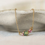A delicate gold pendant necklace displayed on a ceramic dish against a soft cream background, featuring a gemstone cluster of a green square sapphire and a small pink stones with diamonds.