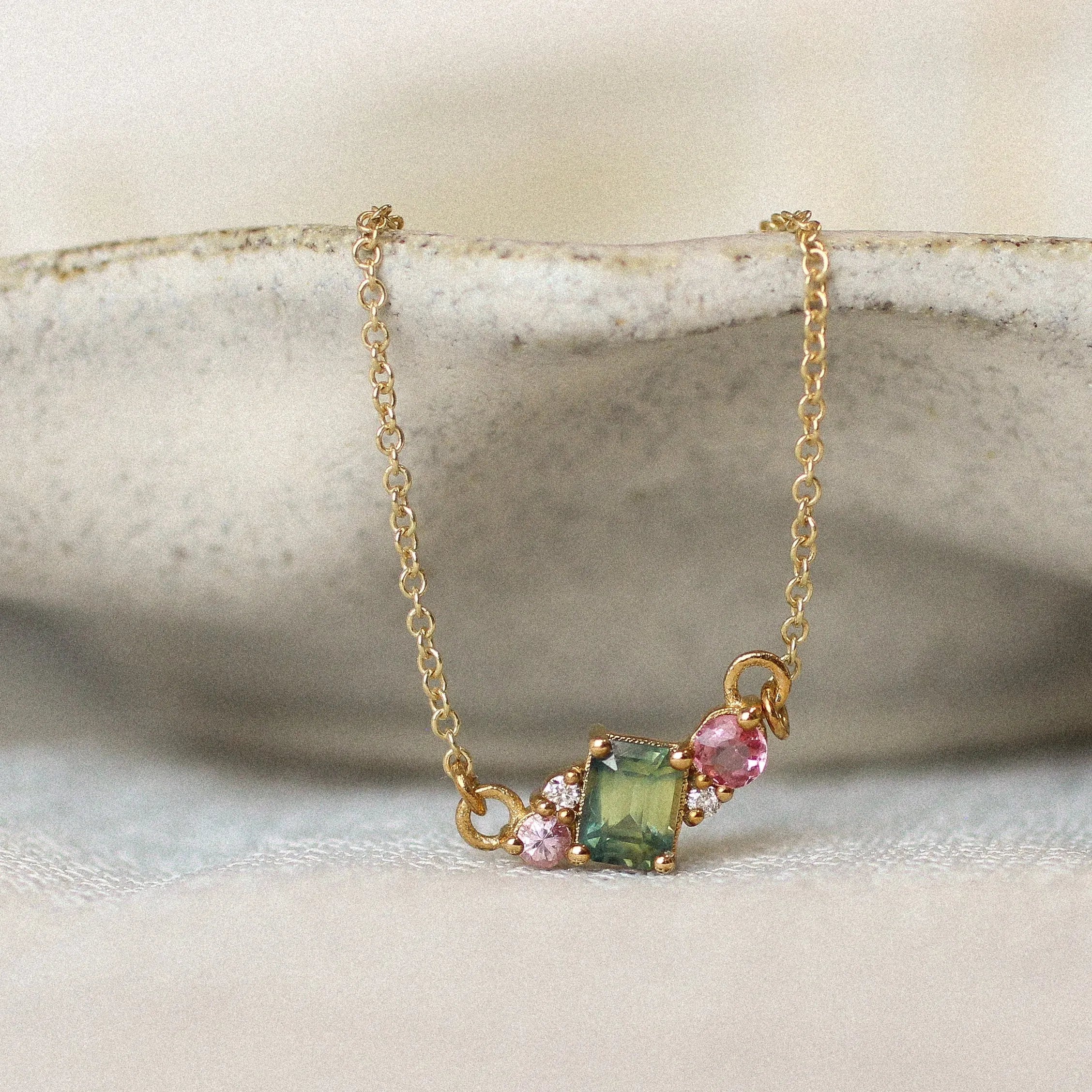A delicate gold pendant necklace displayed on a ceramic dish against a soft cream background, featuring a gemstone cluster of a green square sapphire and a small pink stones with diamonds.