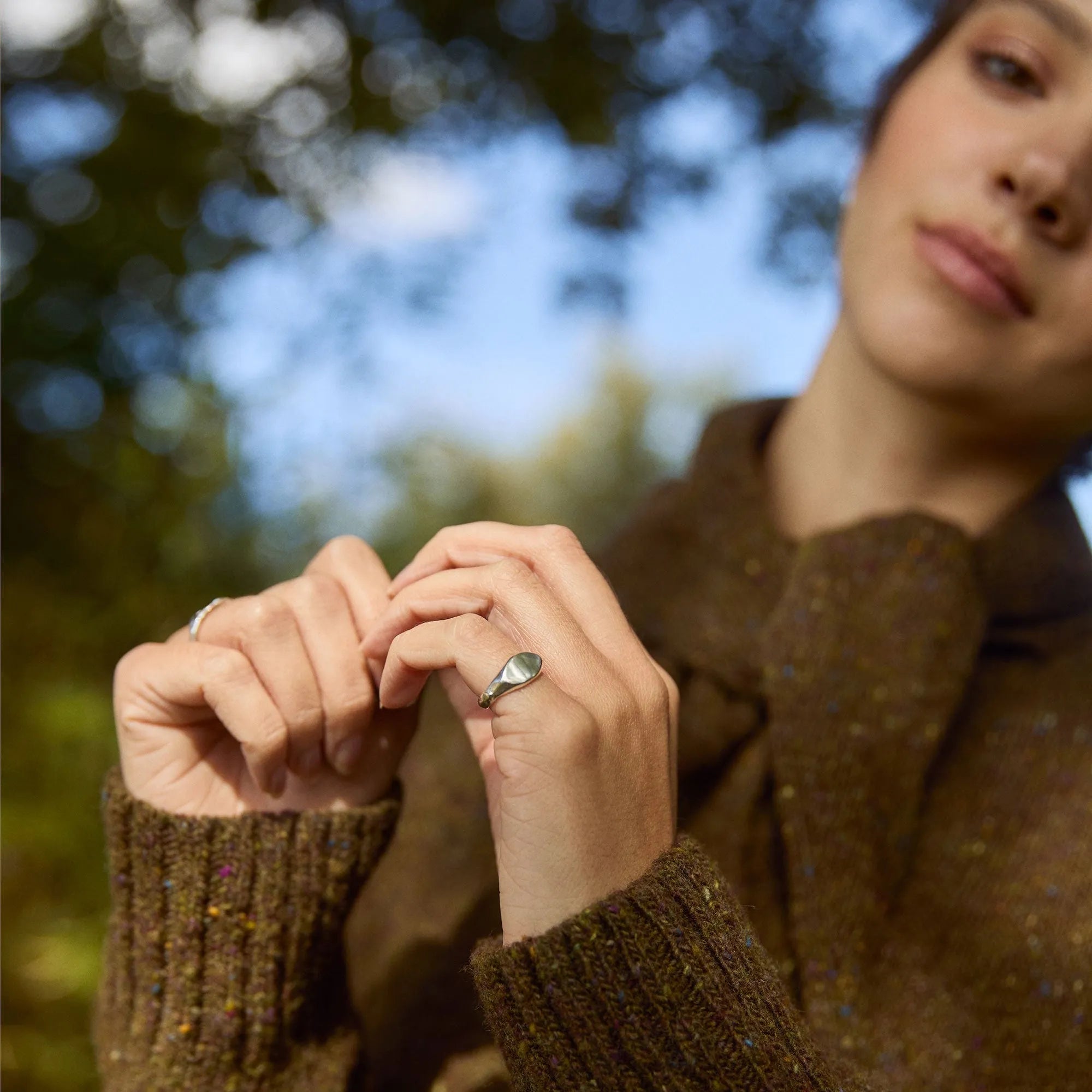 Person wearing a brown sweater and silver pinkie ring with a blurred natural background